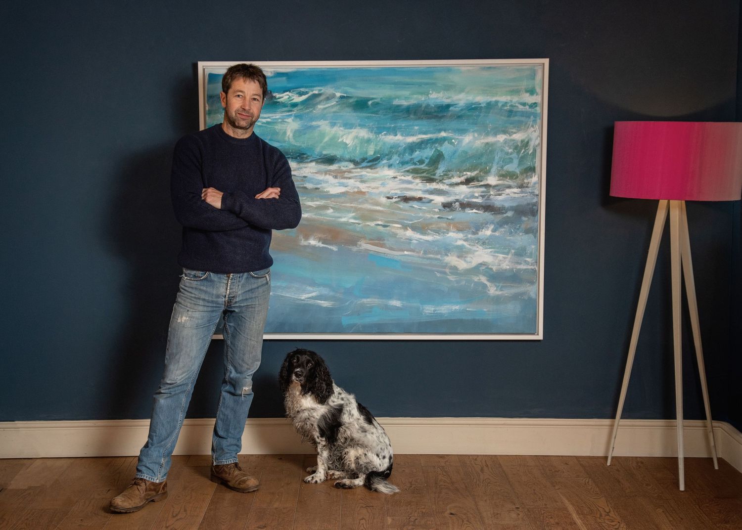 Man in navy sweater and jeans stands beside a spaniel dog in front of a large blue ocean wave painting on a dark wall.