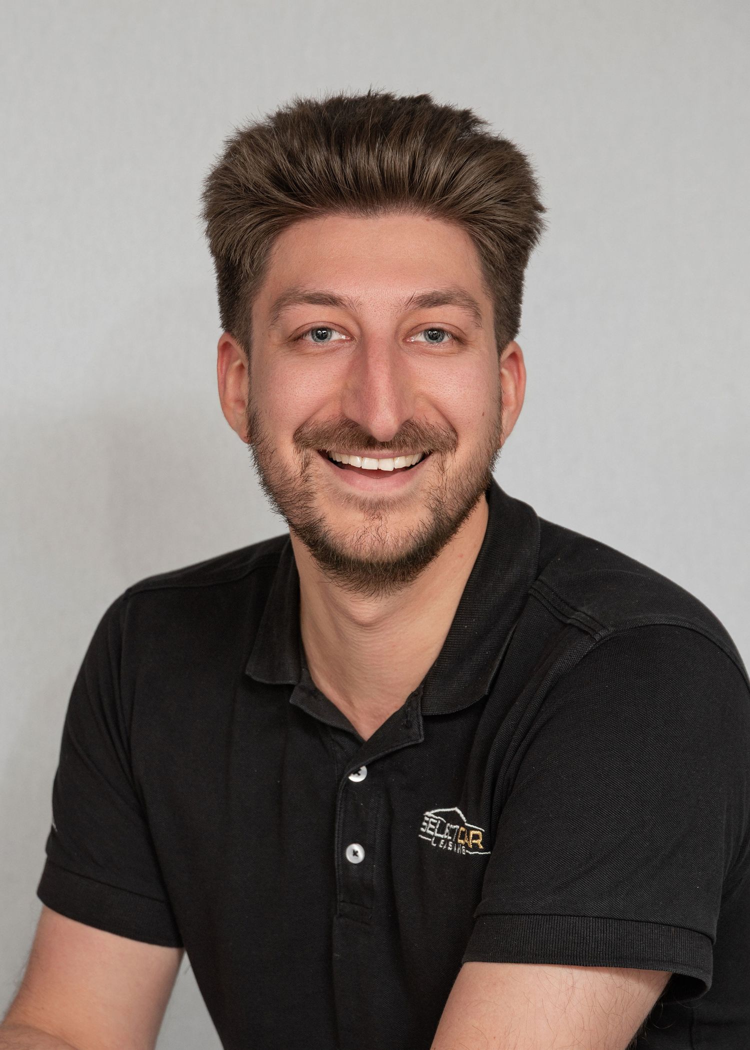Smiling young man with dark hair and beard wearing a black polo shirt with a logo, professional headshot photo.