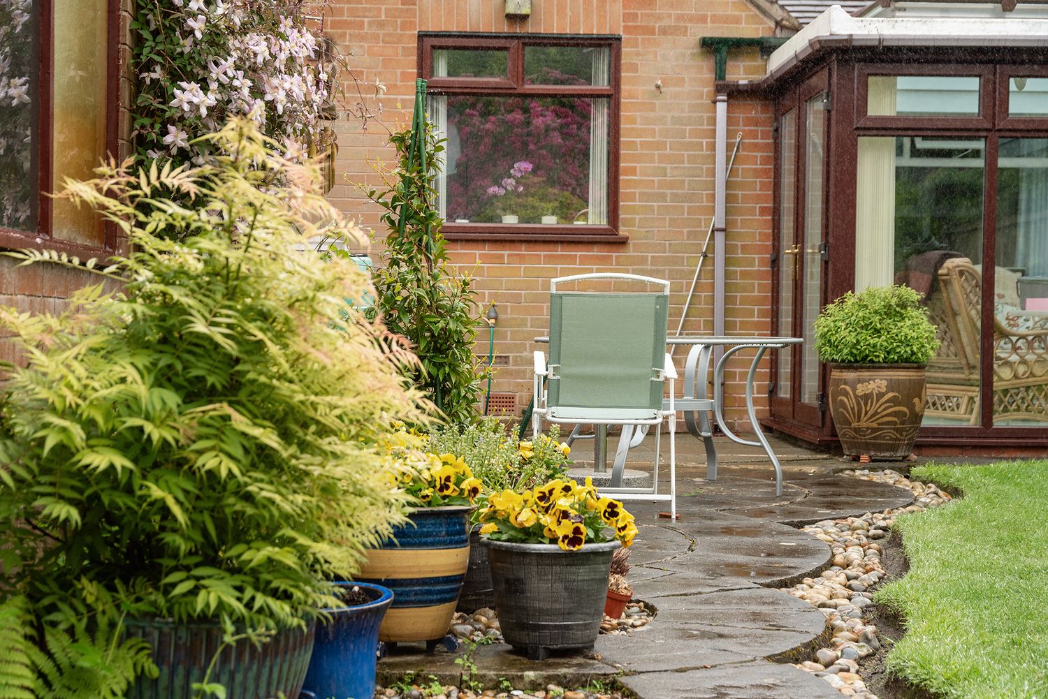 Lush garden patio with potted ferns, yellow flowers, green chair, and brick house conservatory in background.