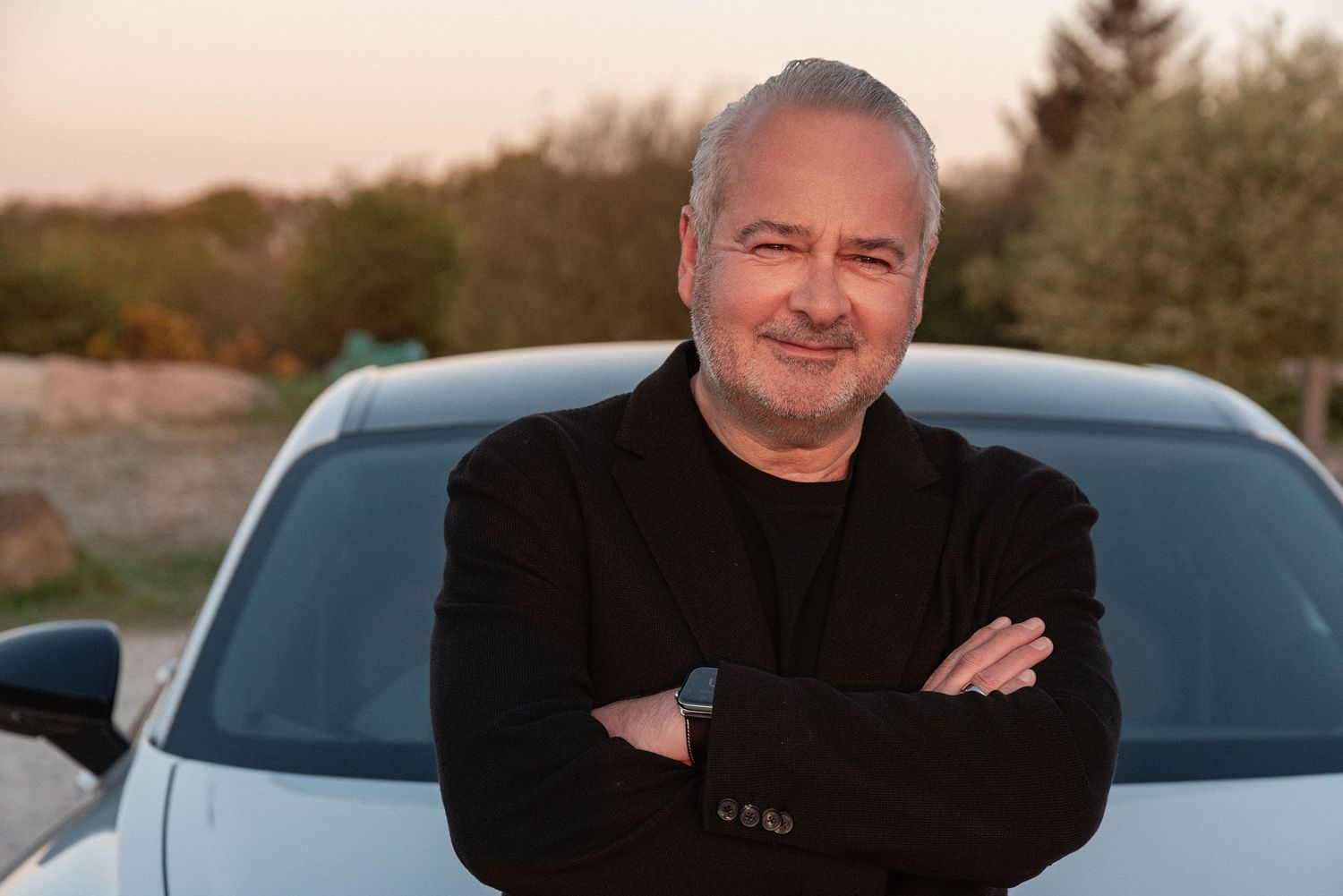 Confident middle-aged man with gray hair, arms crossed, smiling outdoors near a white car at sunset.