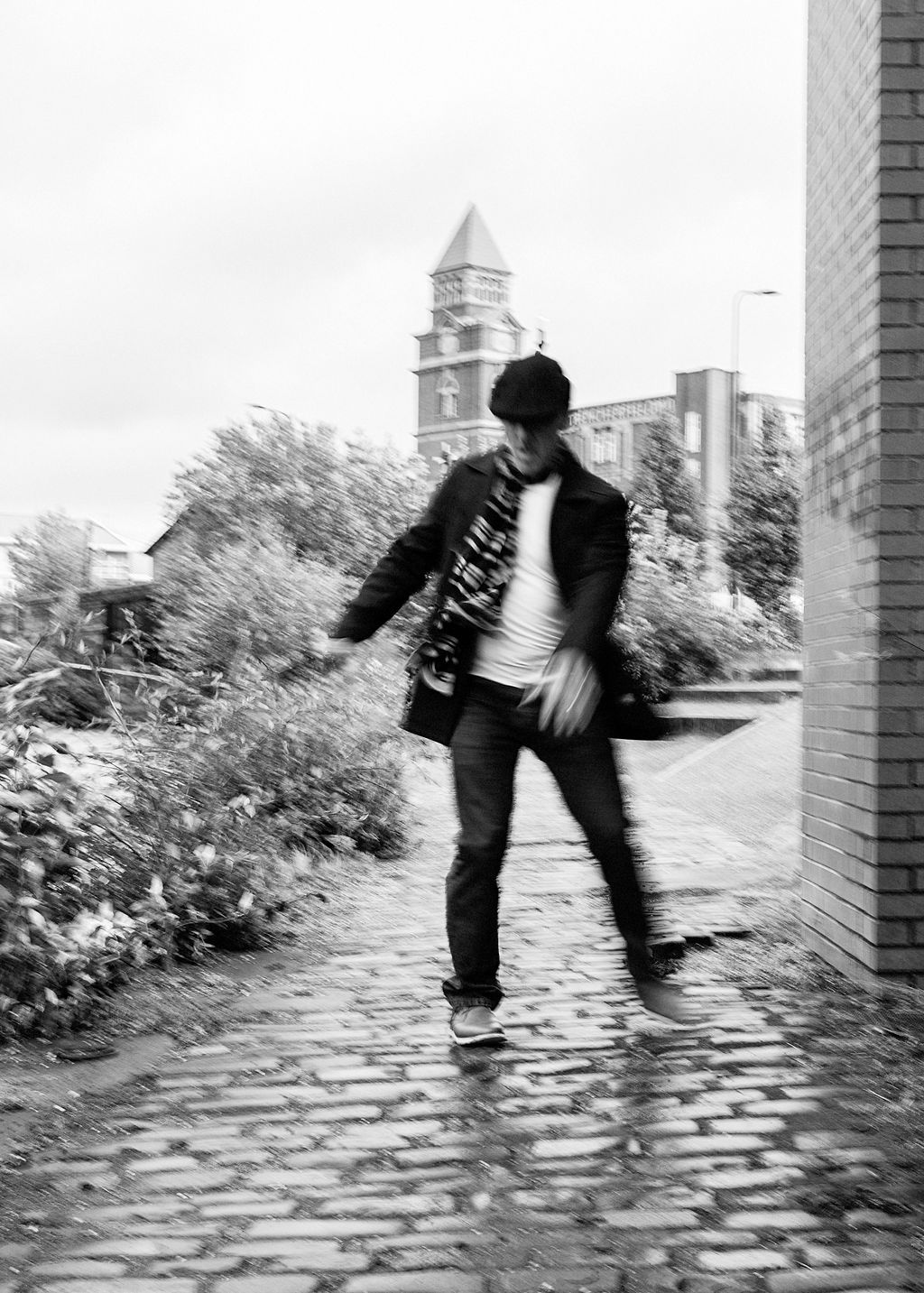 Black and white photo of a man dancing on a cobblestone path near a brick wall with a clock tower in the background.