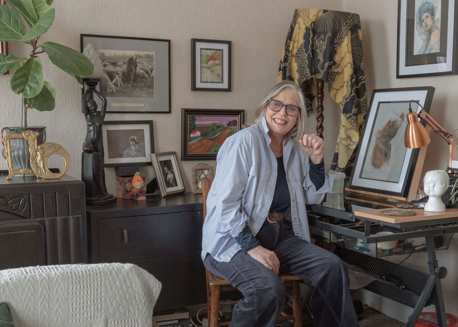 Woman smiling in art-filled home office surrounded by framed artwork, sculptures, and eclectic decor.