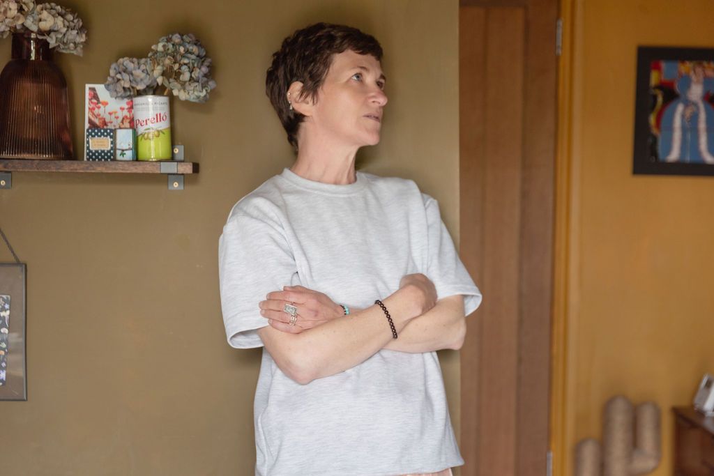 Person with short hair stands indoors with arms crossed, wearing a light gray oversized t-shirt near a shelf with decor.