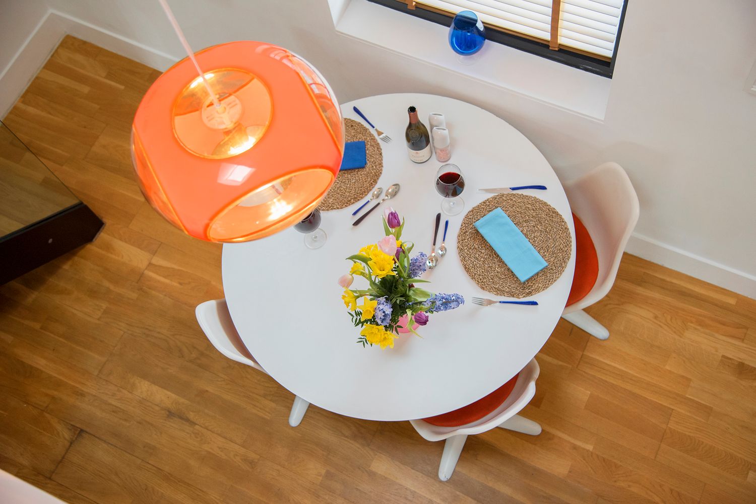 Aerial view of a round white dining table with colorful flowers, blue napkin, and an orange pendant light on hardwood floors.