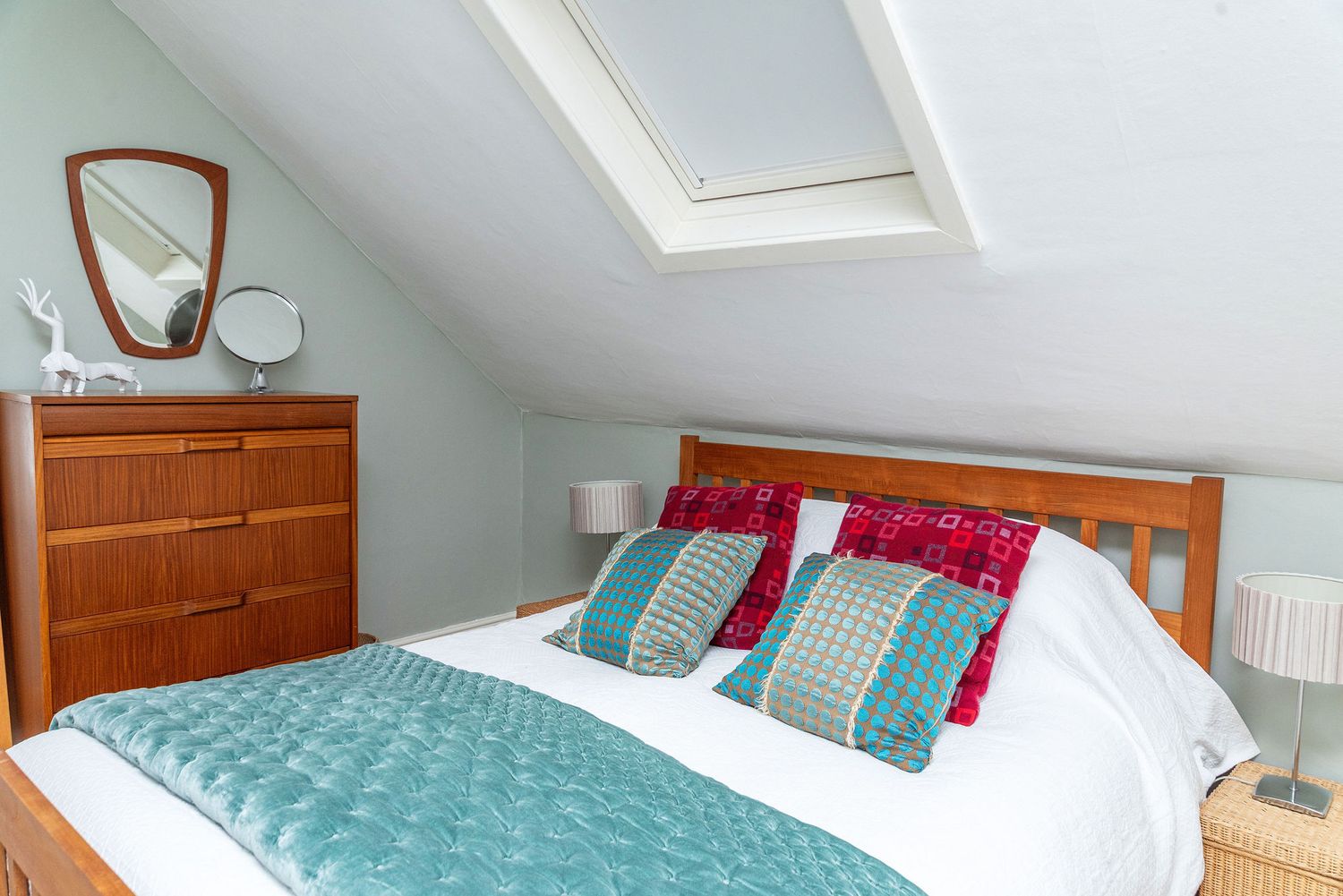 Cozy attic bedroom with skylight featuring teal and red accent pillows on wooden bed frame and matching dresser.