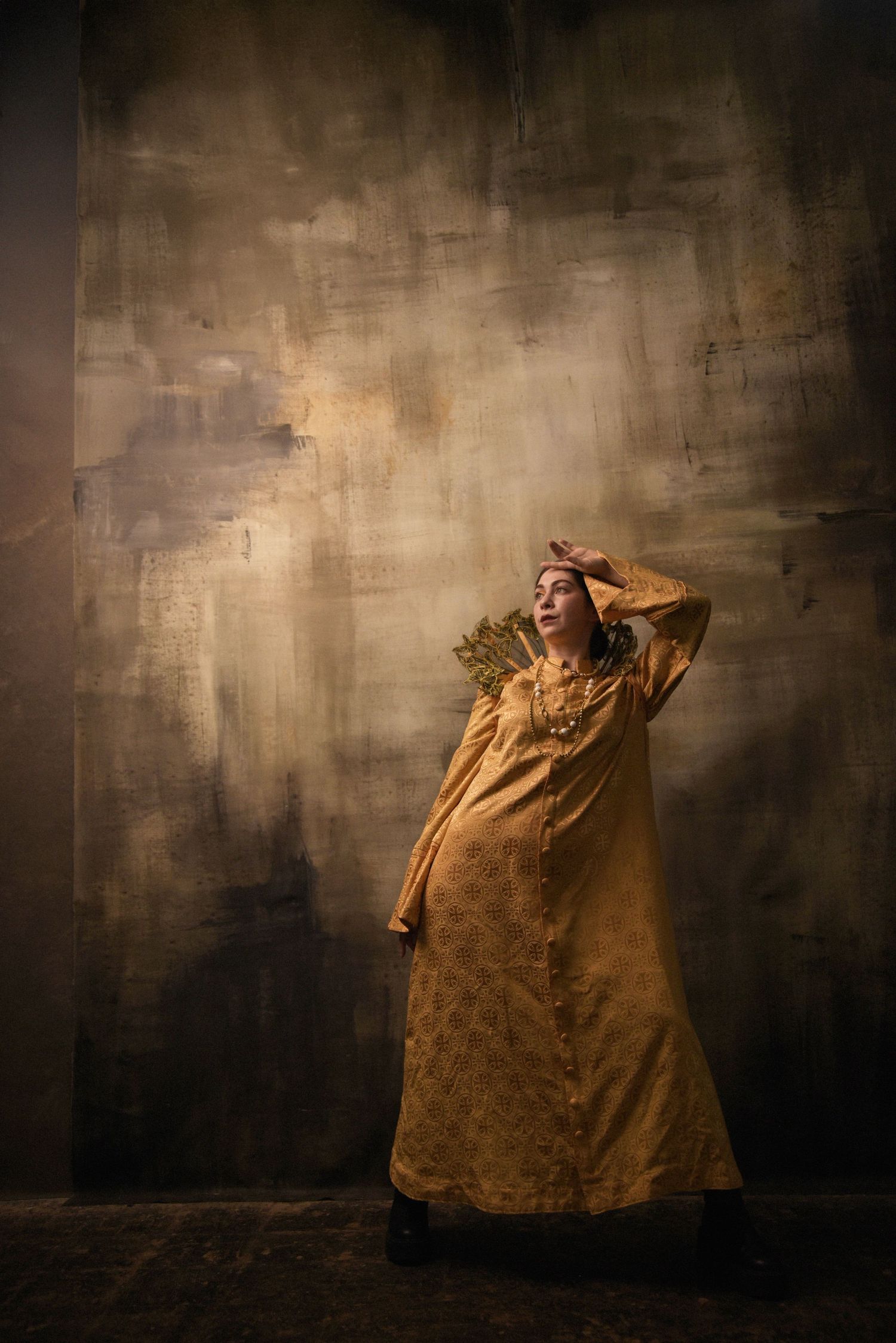 Dramatic portrait in golden dress with theatrical lighting against textured brown wall.