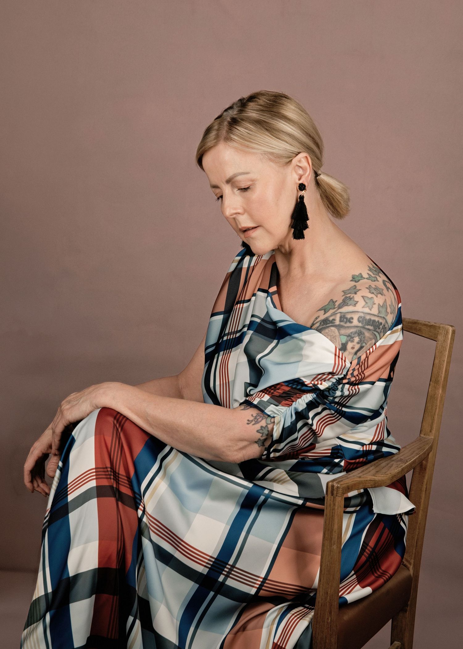 Woman in plaid dress seated on wooden chair, wearing black tassel earrings, with tattoos visible, posed against neutral background.
