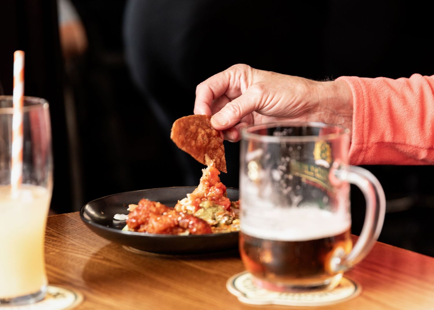 Person dipping a chip into a spicy appetizer on a dark plate, with a beer mug and juice glass on a wooden table.