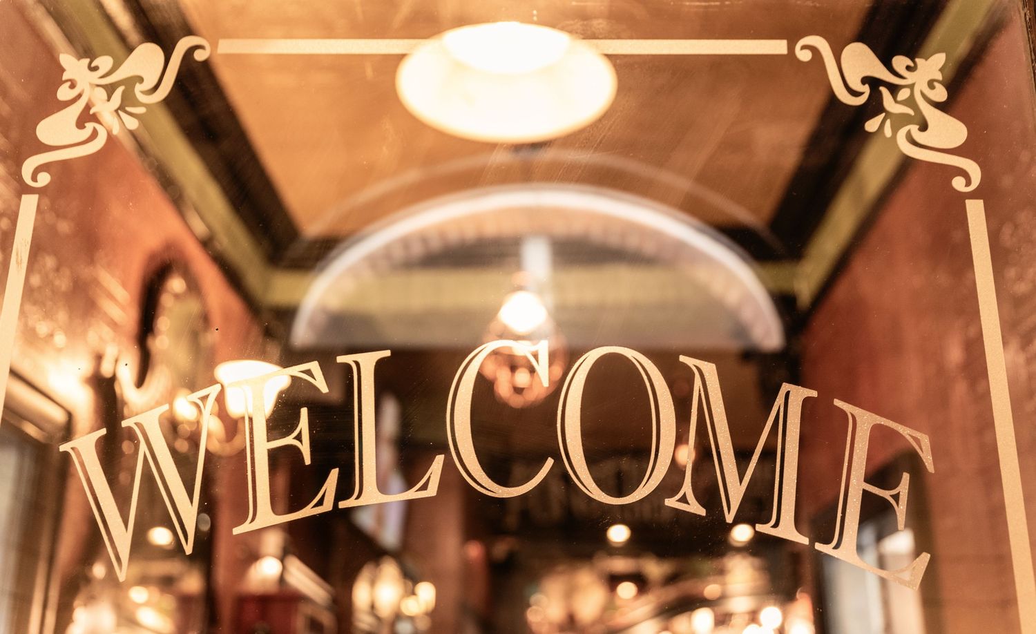 Golden decorative WELCOME sign displayed on glass door of elegant vintage restaurant interior.