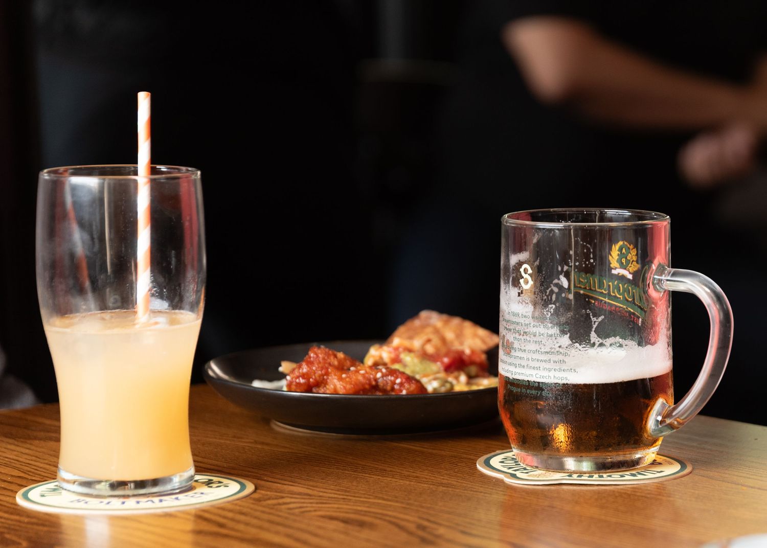 Two drinks on a bar, a fruity cocktail with straw and a beer mug, alongside a plate of chicken wings.