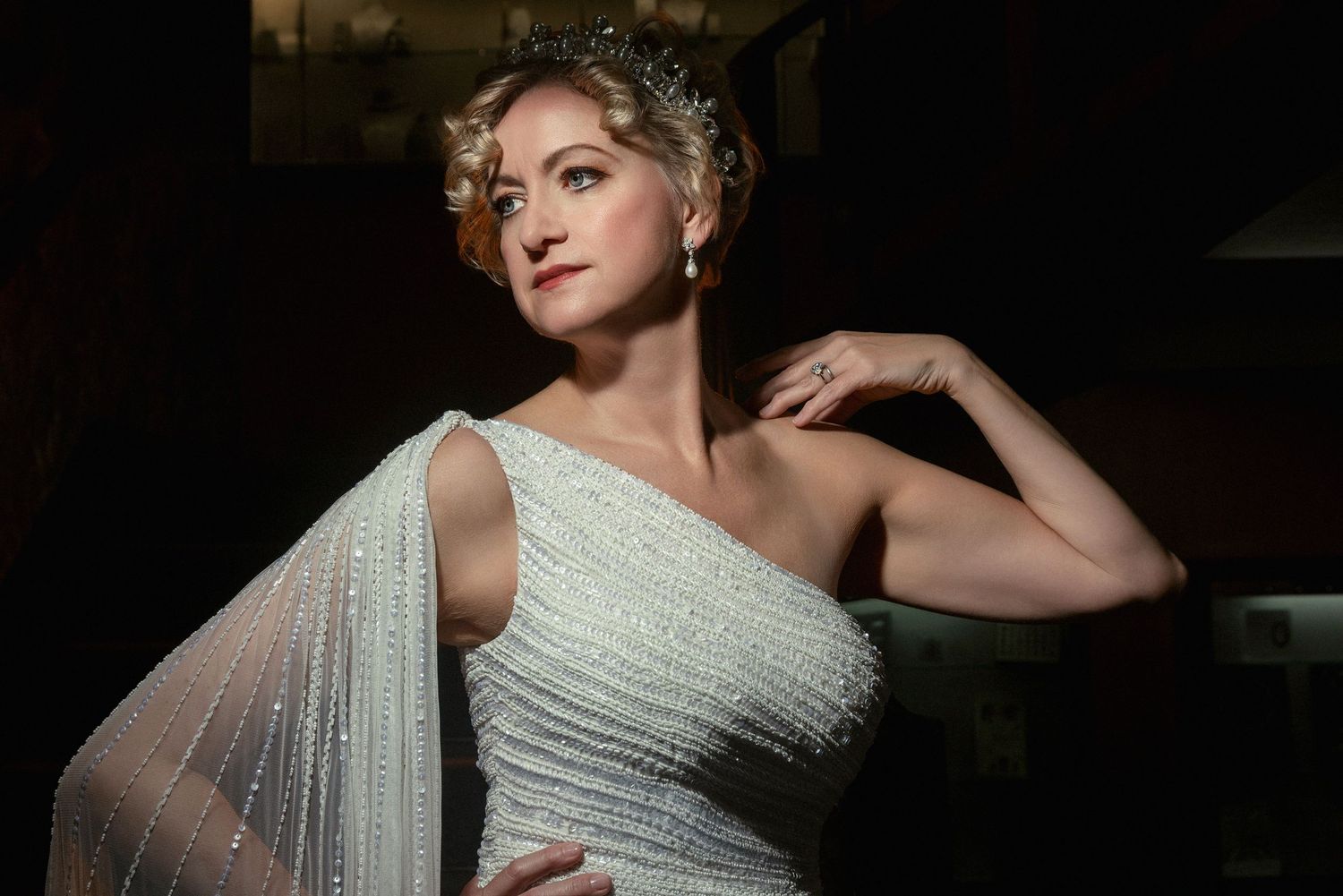 Elegant woman in a sparkling white one-shoulder gown and crystal tiara posing gracefully in dramatic lighting.