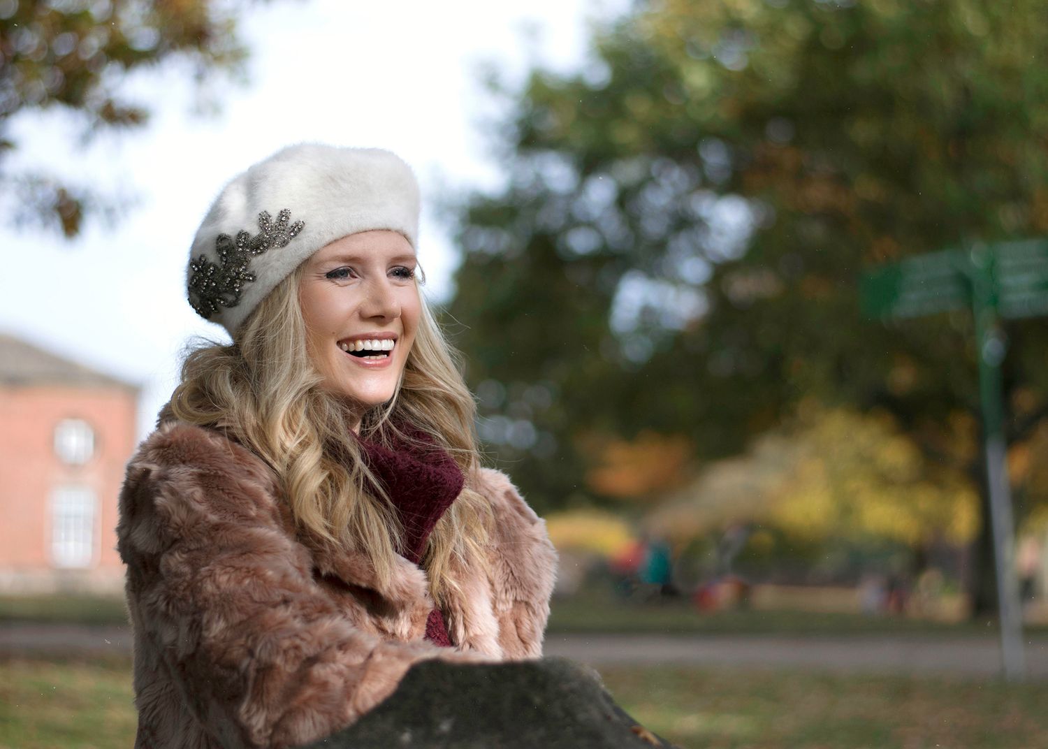 Smiling blonde woman wearing a white beret and fur coat outdoors in autumn park setting.