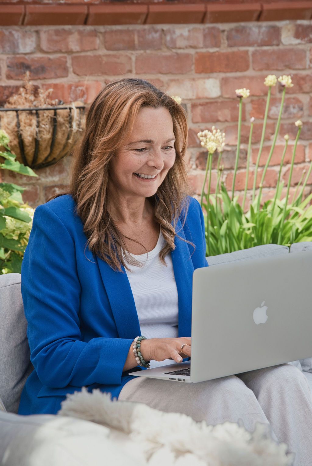 Woman in blue blazer smiling while working on Apple MacBook laptop outdoors in garden setting.