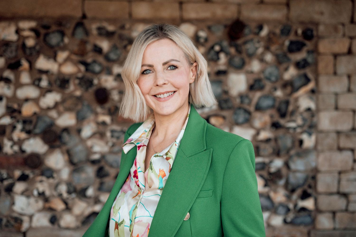 Smiling blonde woman in green blazer and floral blouse posing outdoors against a rustic stacked wood wall.