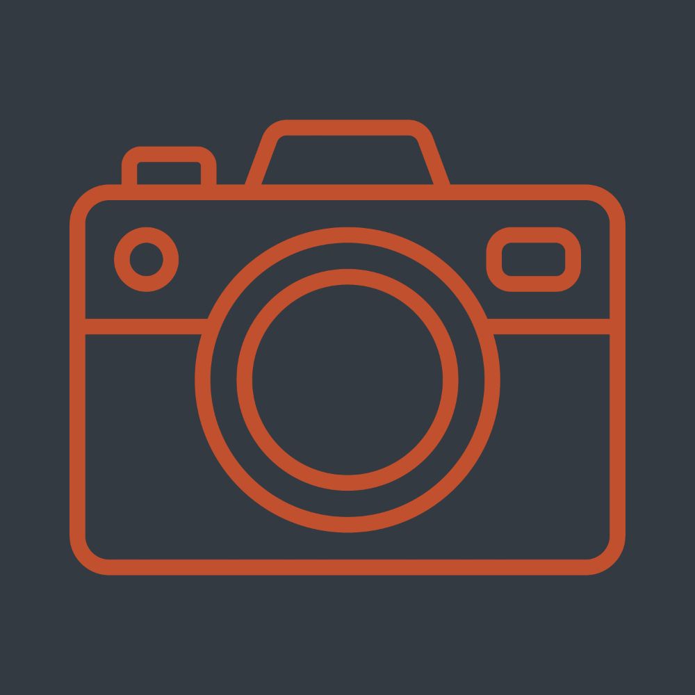 Orange line-art icon of a digital camera with a round lens, flash, and viewfinder on a dark charcoal background.