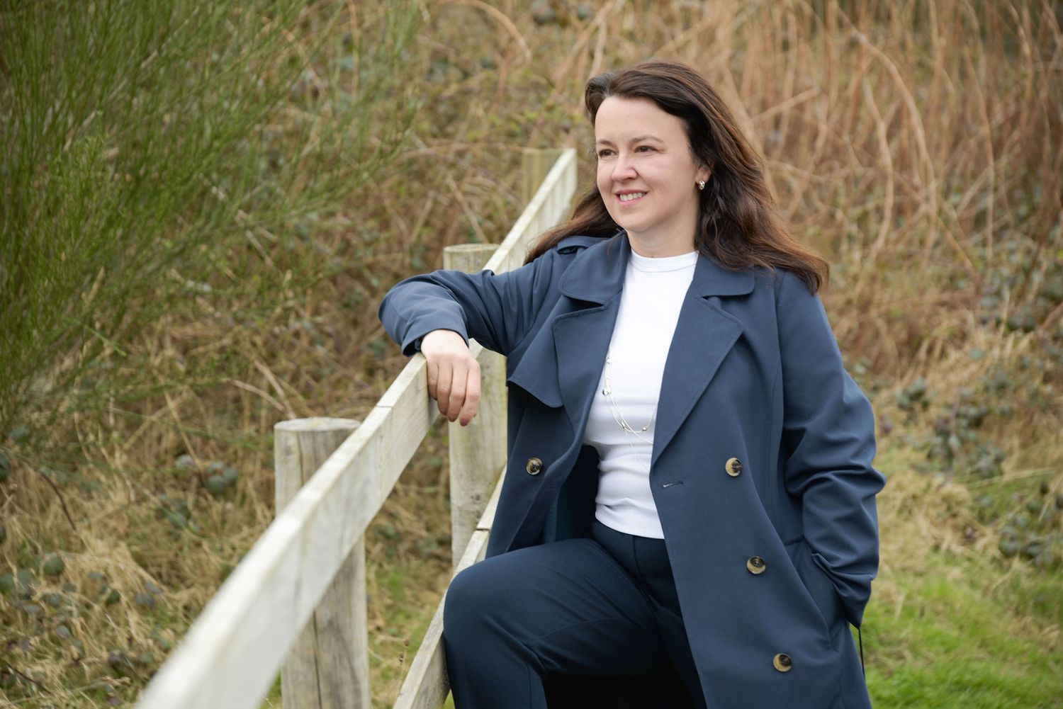 Someone in a navy blazer and white shirt leans casually against a wooden railing in a natural outdoor setting.