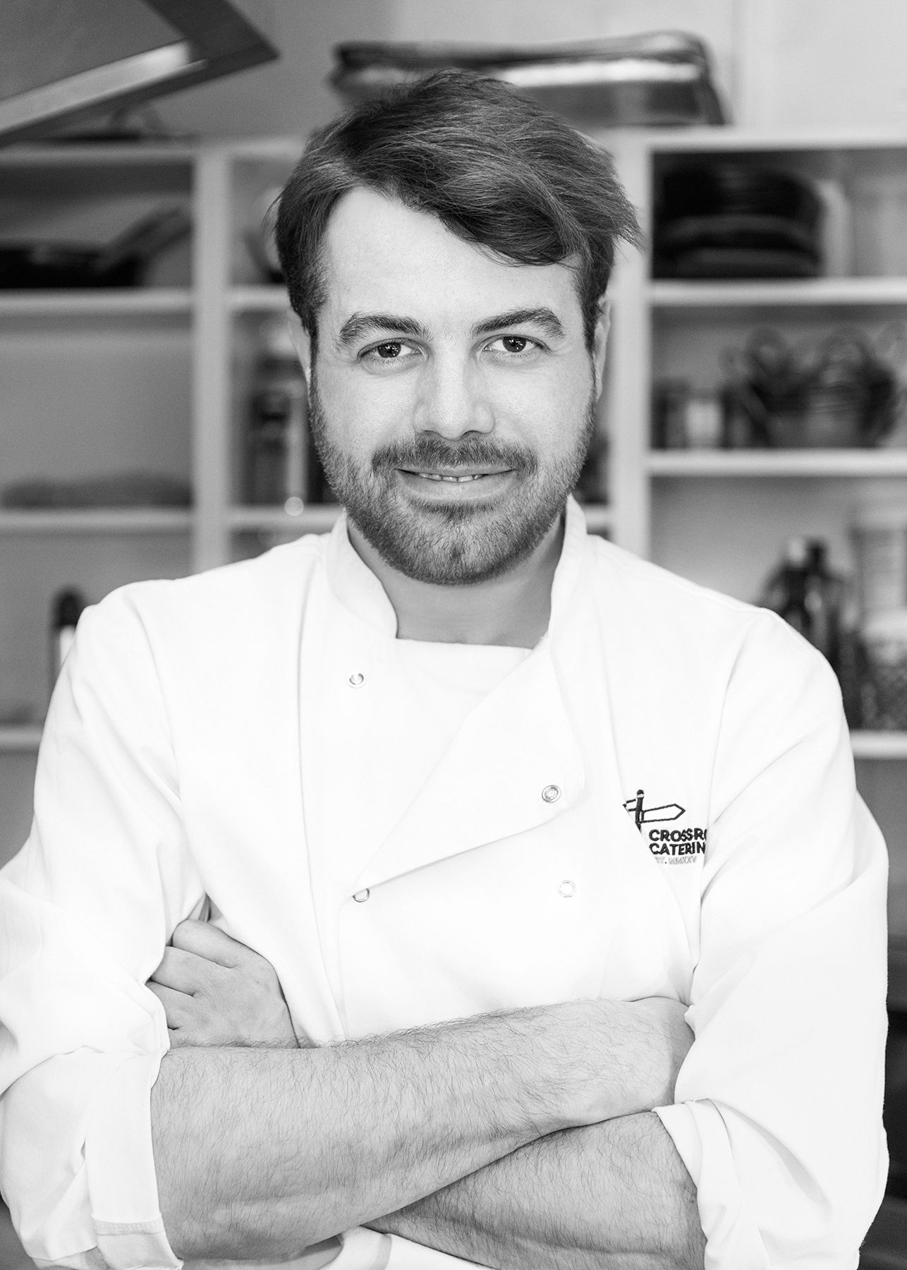 Professional chef in white uniform smiling with arms crossed in a modern kitchen, black and white portrait photo.