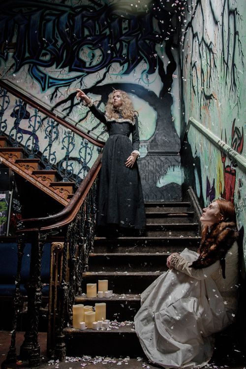 Two women in gothic Victorian gowns pose on a graffiti-covered staircase with candles in a dark setting.
