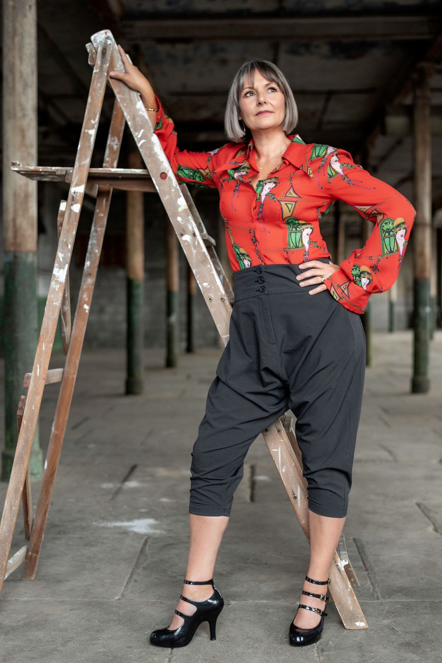 Fashion model in red floral blouse and dark culottes poses elegantly near wooden ladder in industrial setting.
