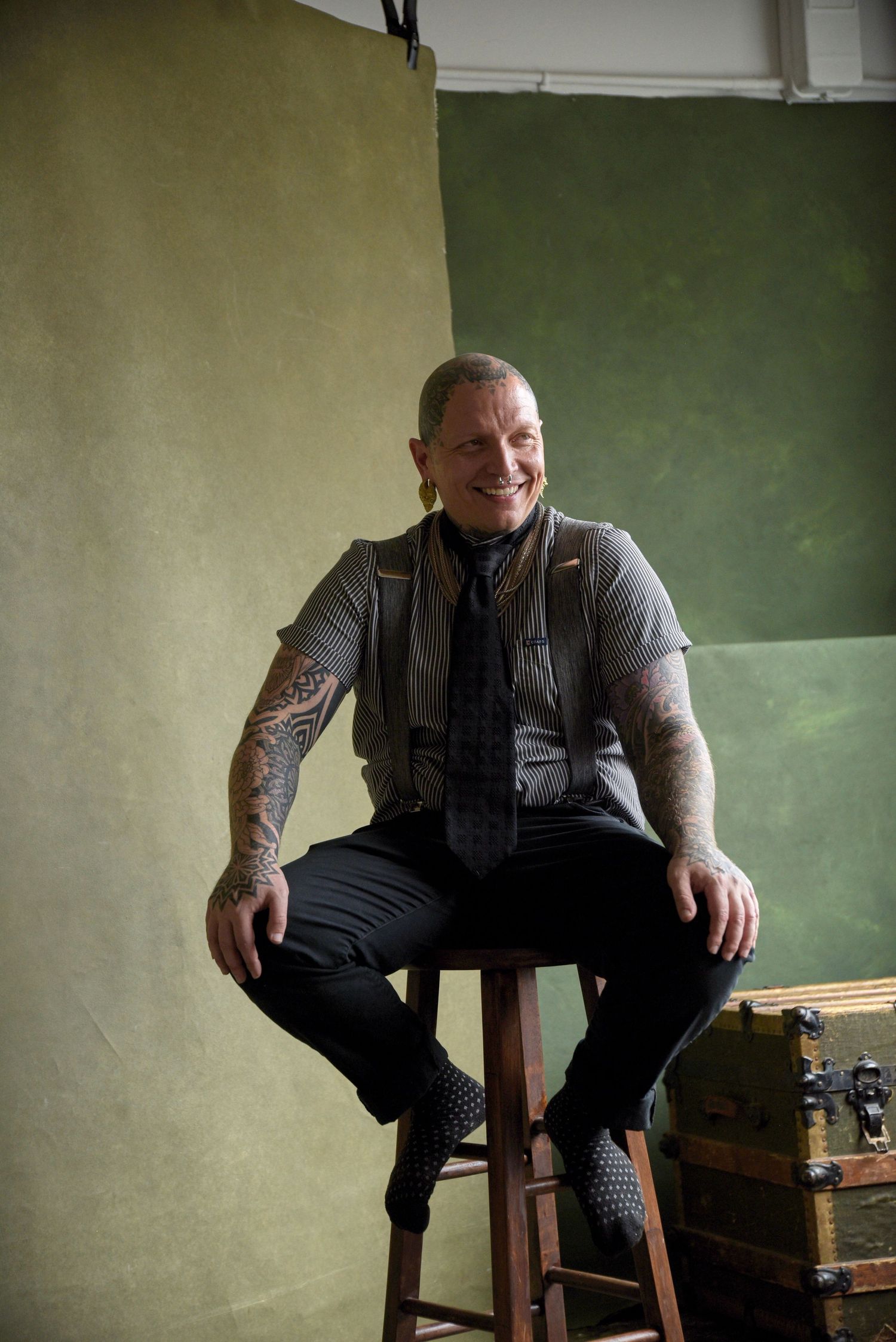 Tattooed model in business attire sits on stool for studio portrait.