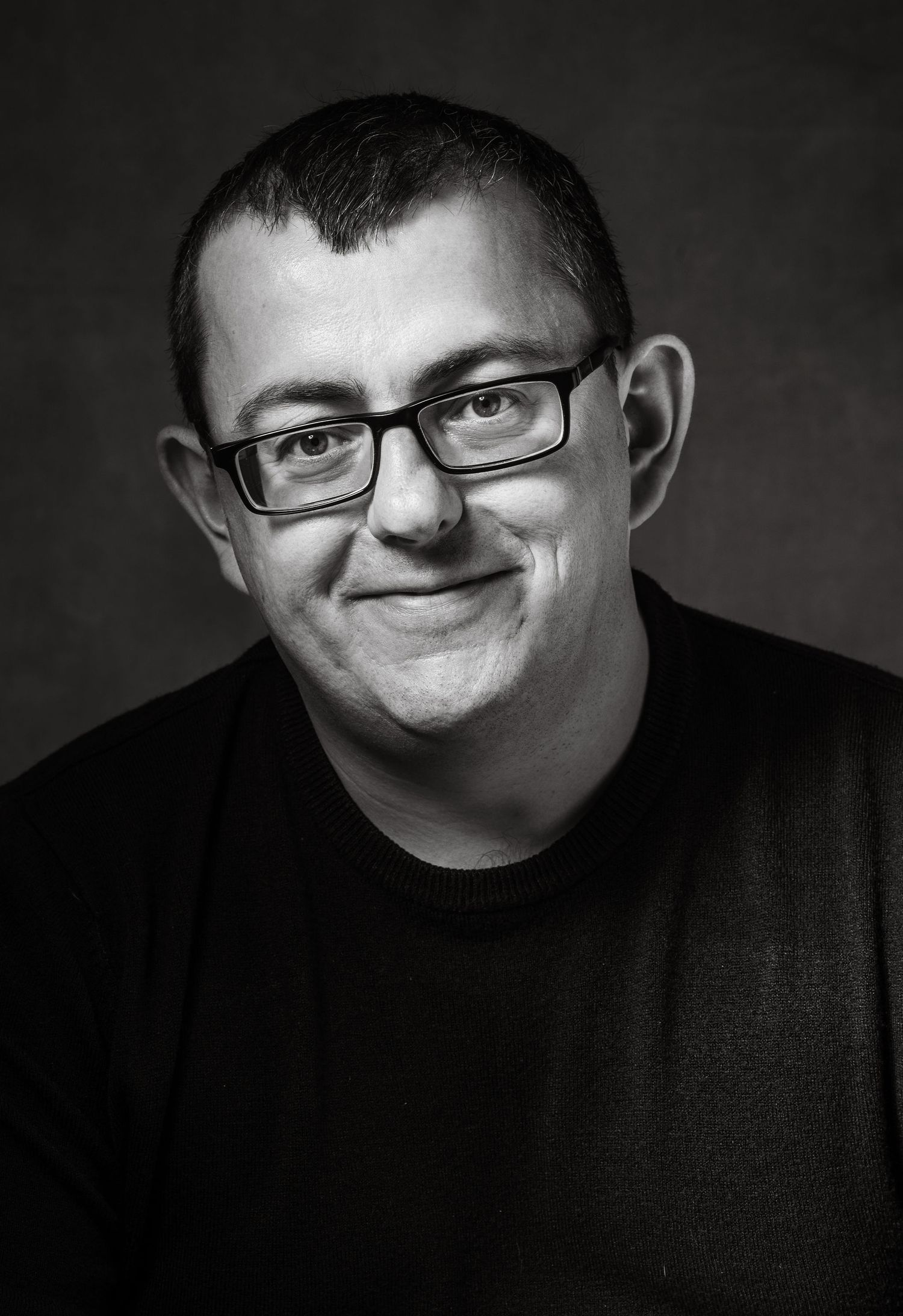 Black and white headshot portrait of person wearing glasses and dark shirt against dark background.