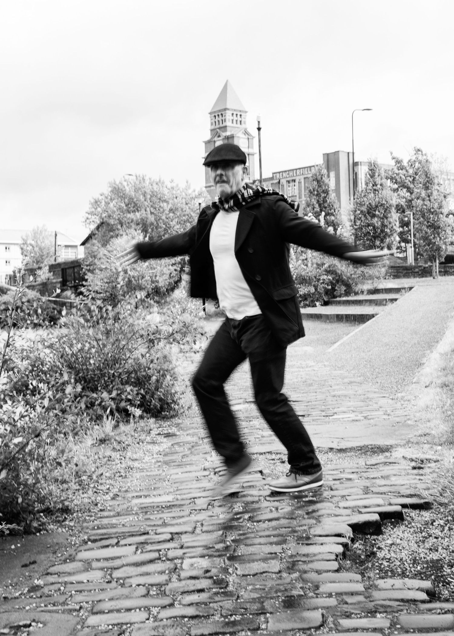 A person in a formal suit performs a playful dance move on a cobblestone path with a church spire visible in the background.