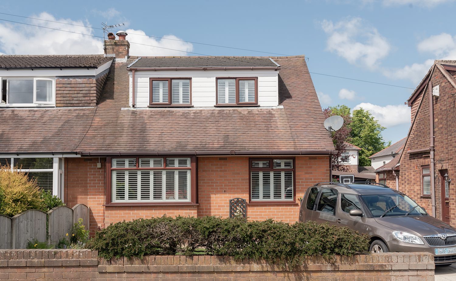 Semi-detached brick house with dormer windows, satellite dish, and a car parked on the driveway behind a low fence.