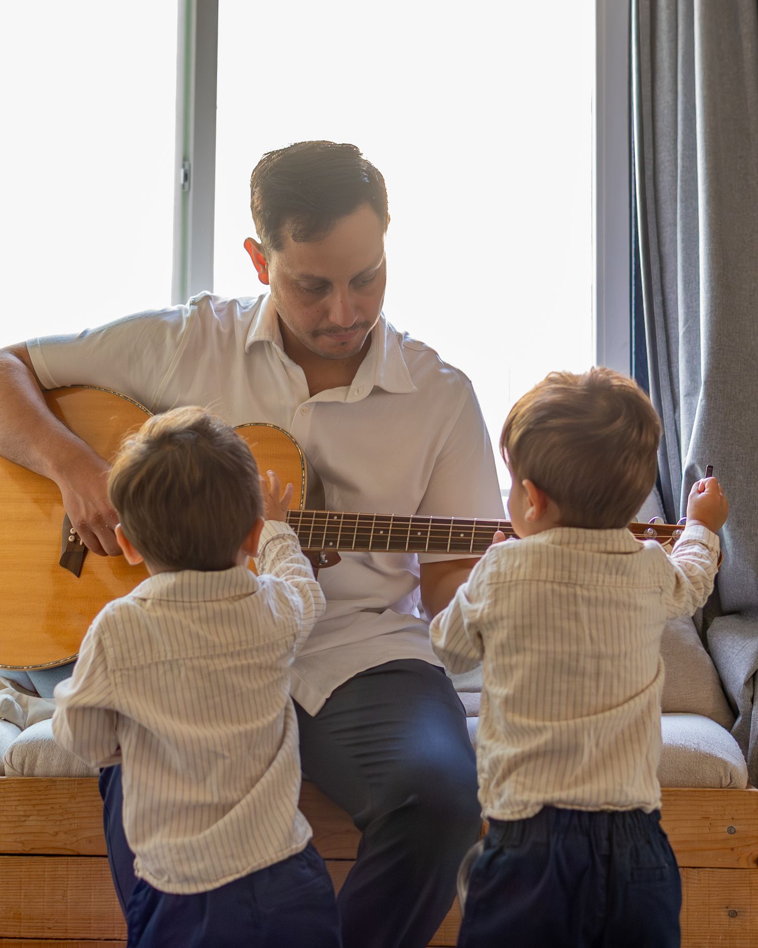 A father plays acoustic guitar for his two toddler sons in matching striped shirts by a bright window.