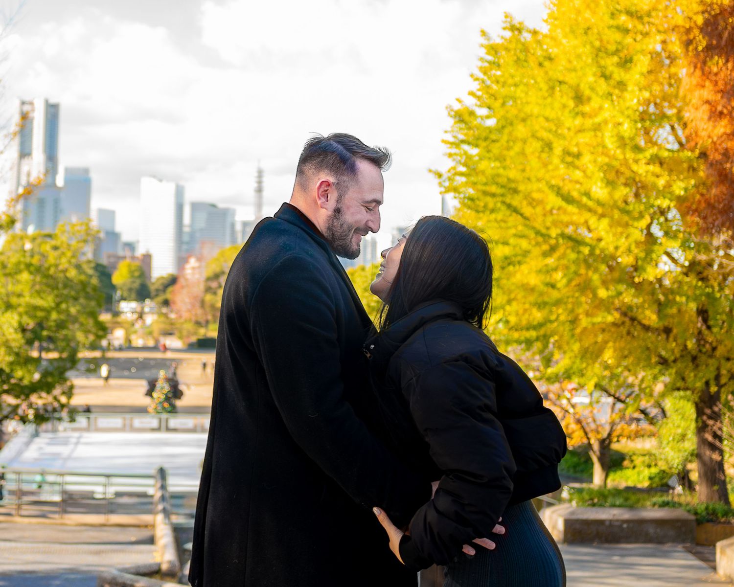 A couple in dark coats embrace in front of vibrant yellow autumn trees with a city skyline in the background.