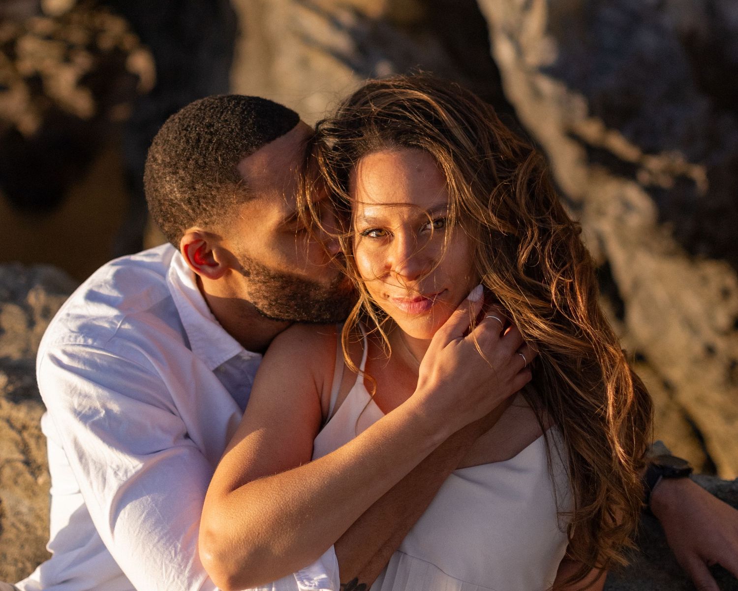 Romantic couple embraces during golden hour photoshoot on rocky beach with warm sunset lighting.