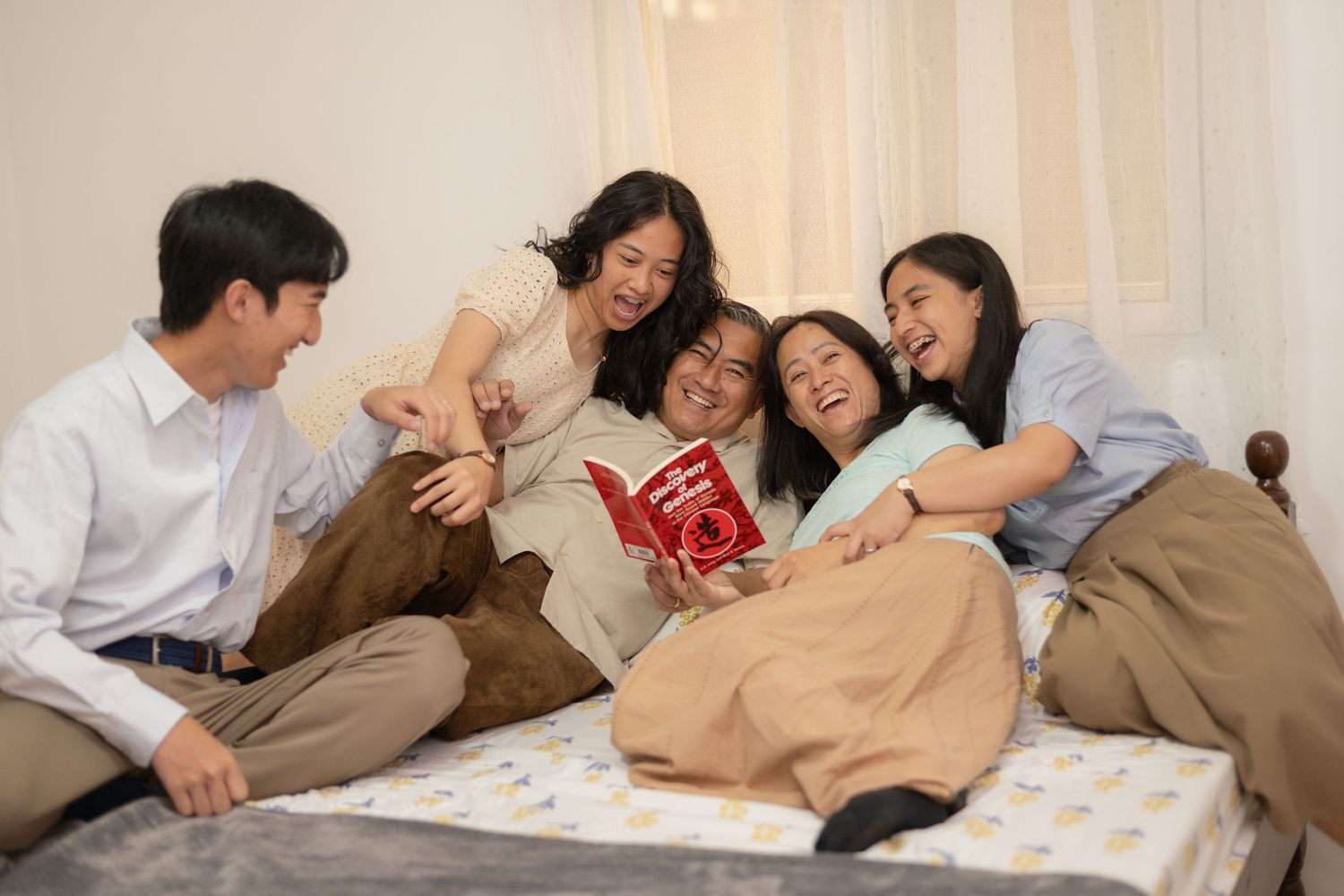 Asian family laughing together on bed while reading a red book, sharing a joyful bonding moment at home.