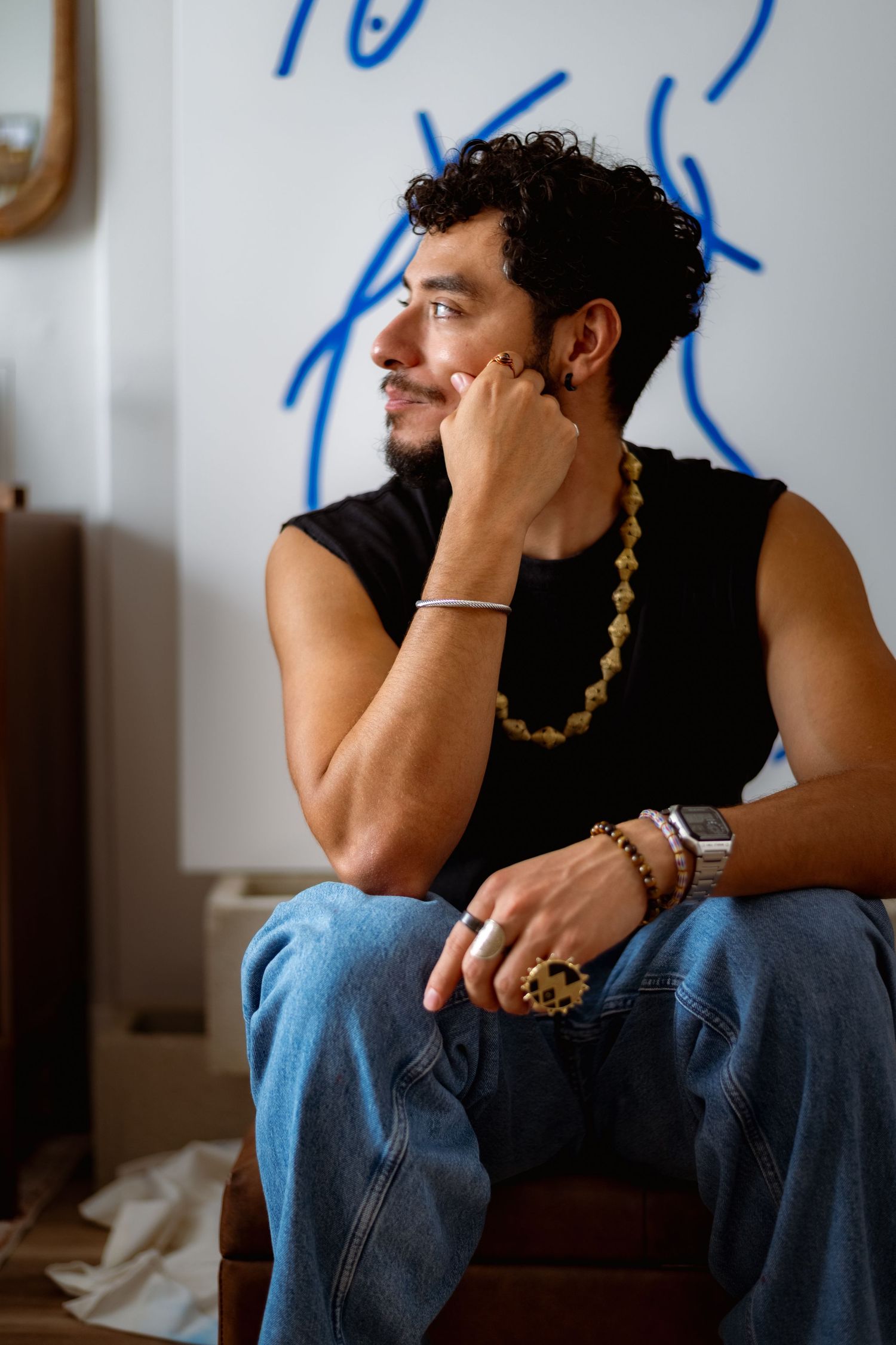 Person in black tank top and jeans sits pensively with hand on face in a dimly lit room.