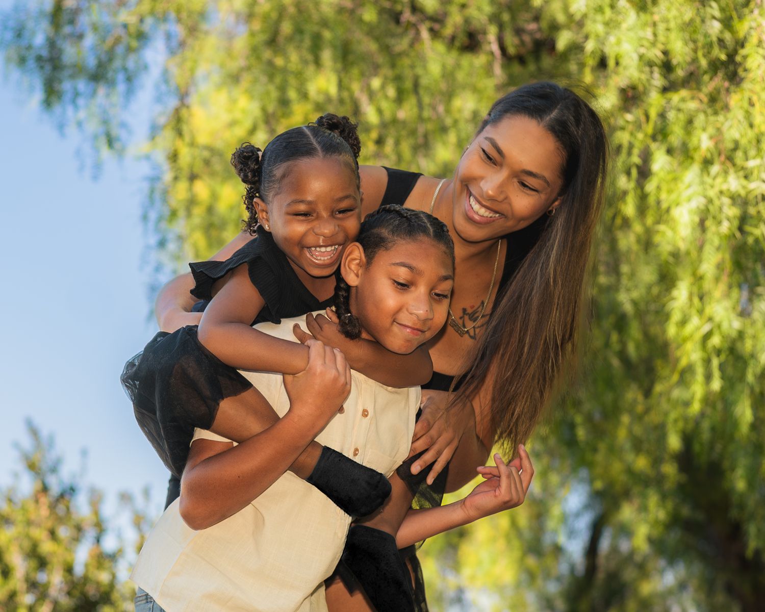Family members share joyful embraces and laughter in a sunny outdoor setting with willow trees in the background.