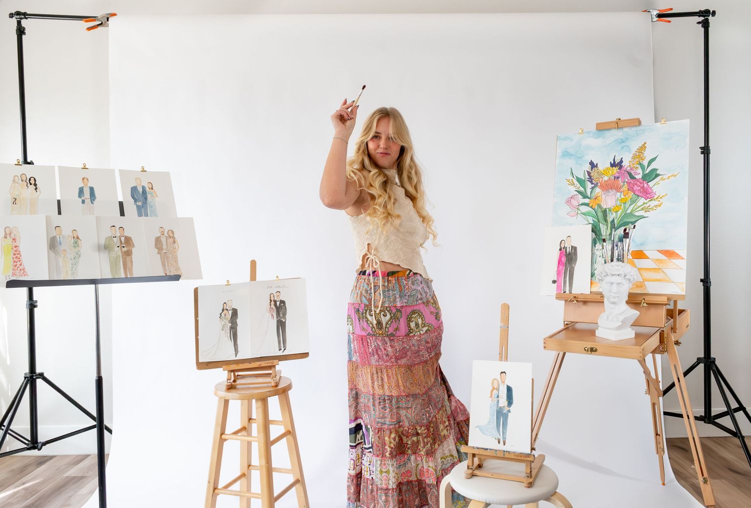 Artist in bohemian skirt poses with paintbrush in bright studio surrounded by fashion sketches and floral paintings.
