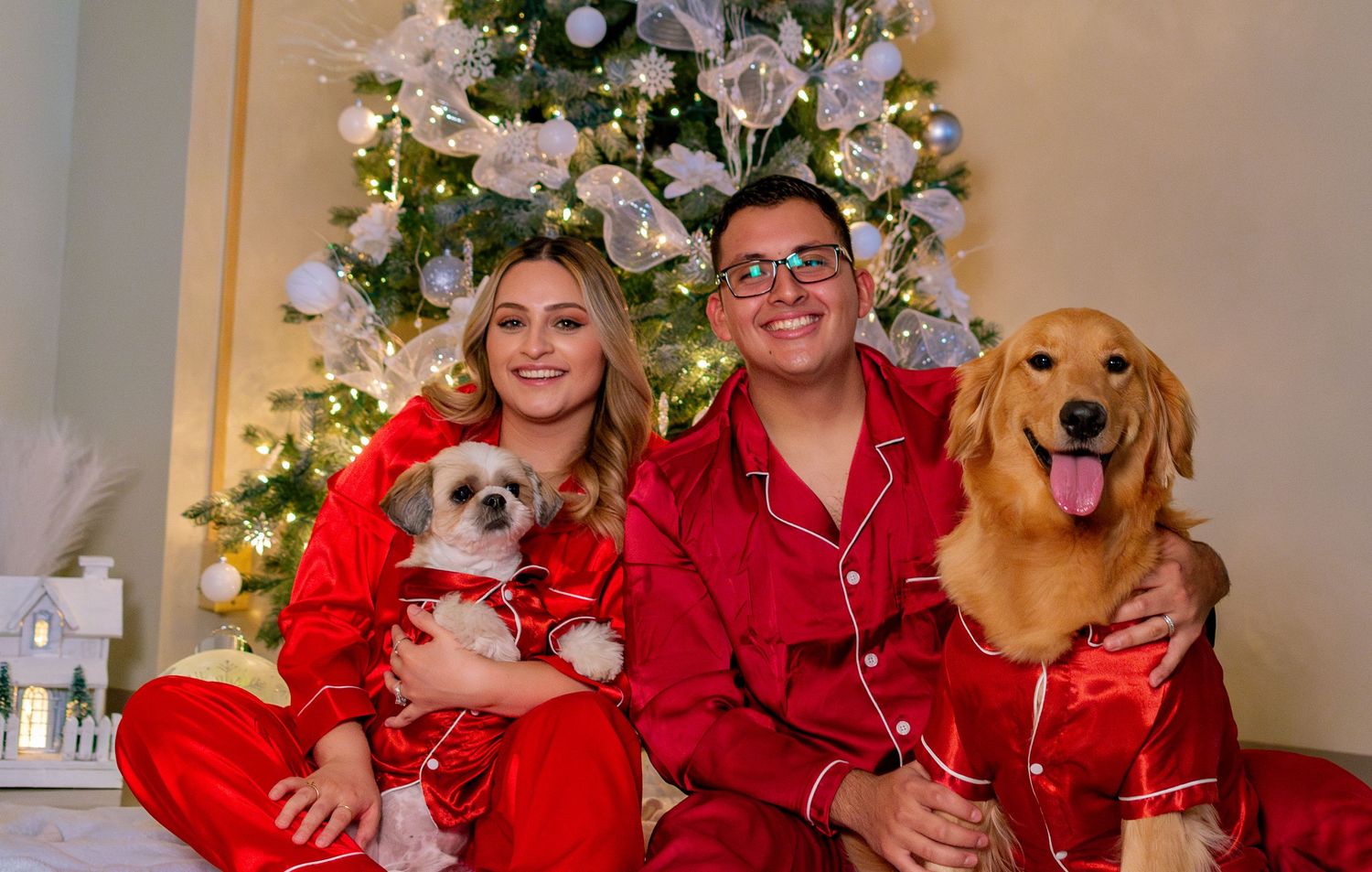 Couple in matching red Christmas pajamas sitting with two dogs in front of decorated holiday tree.