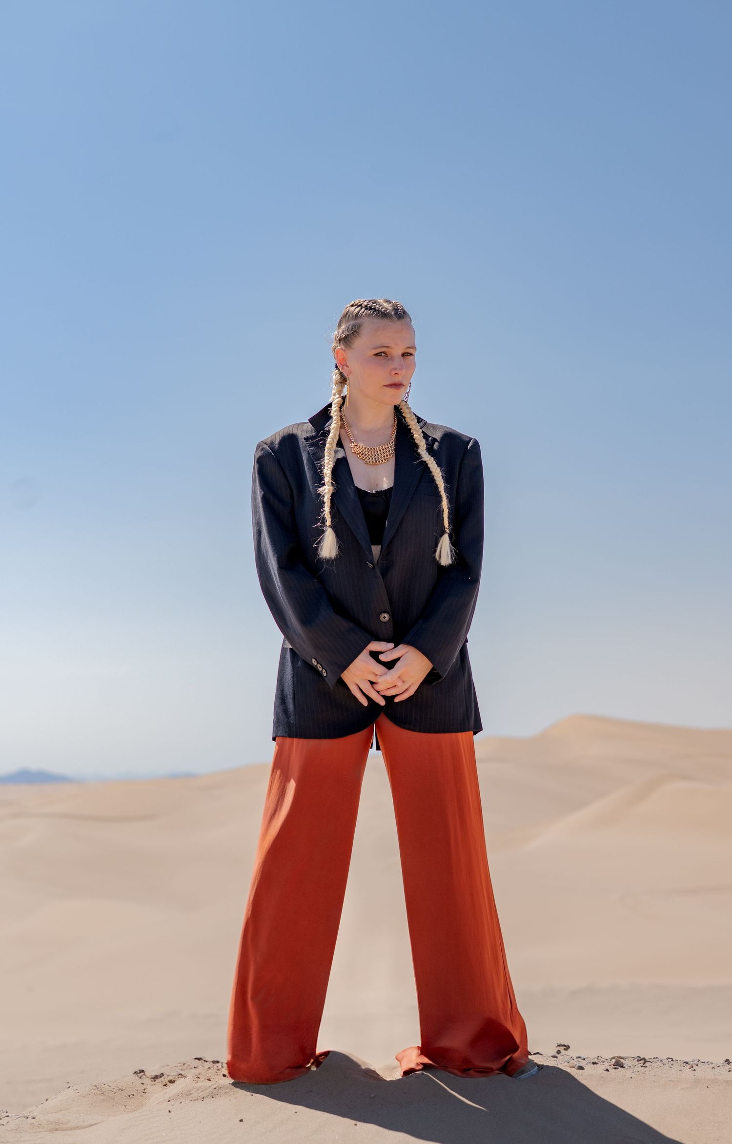 Fashion model poses in black jacket and orange pants against desert sand dunes in dramatic photoshoot series.