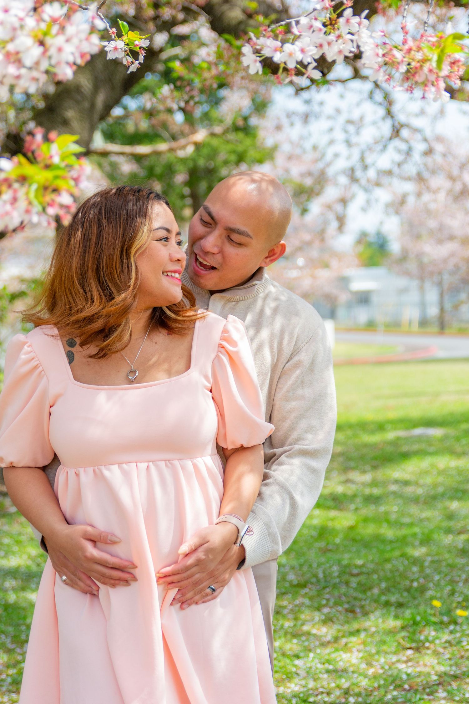 A couple embraces under blooming cherry blossoms in a spring park wearing pink and white outfits.