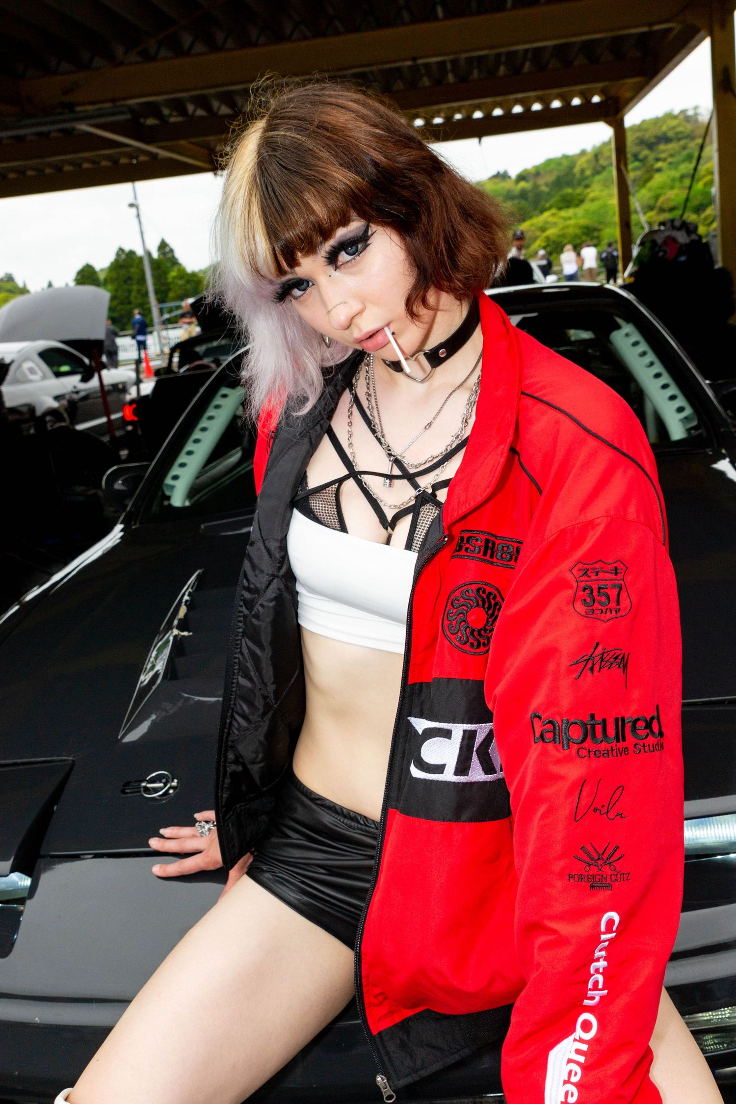 Person wearing red and black streetwear poses with luxury car in parking garage.