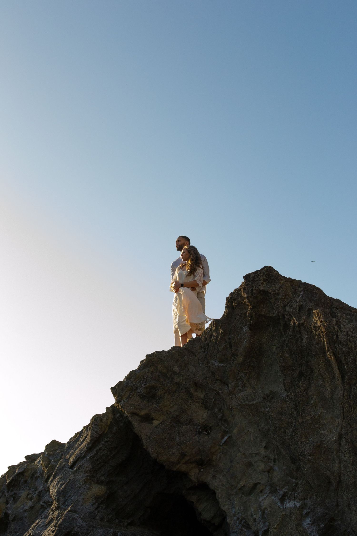 A person stands atop a rocky mountain cliff against a blue sky in a scenic landscape view.