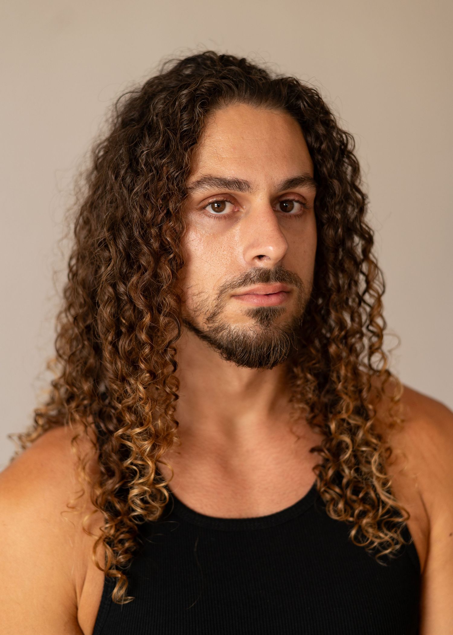 A person with long curly brown hair and a beard poses in a black tank top against a beige background.