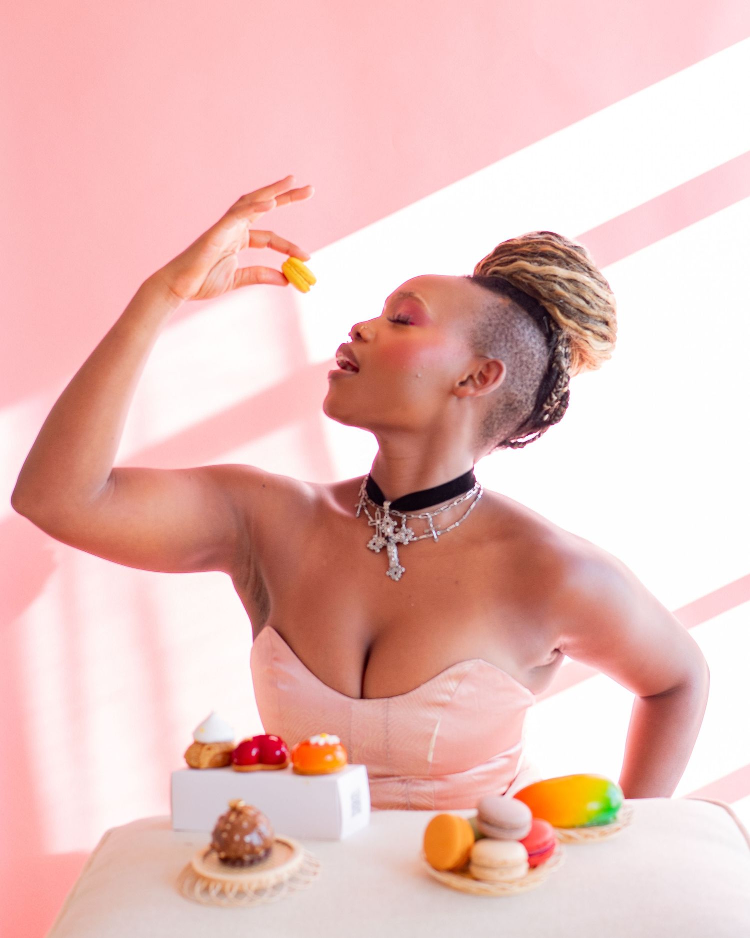 Artistic portrait showing someone enjoying colorful macarons and fruit against a pink backdrop with diagonal light beams.