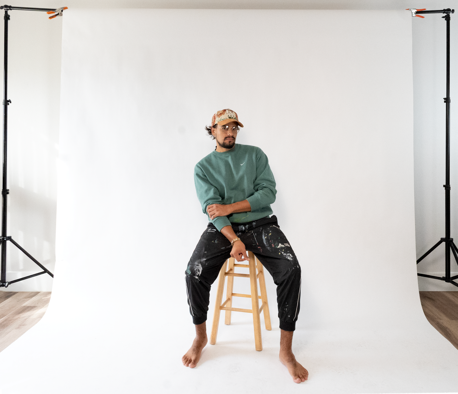 A person in a teal sweater and dark pants sits casually on a wooden stool against a white studio backdrop.