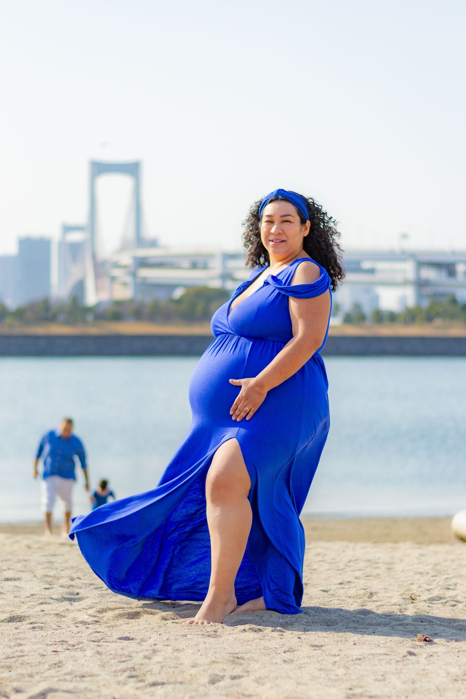 A person in a flowing royal blue dress poses gracefully on a sandy beach with water and buildings in the background.