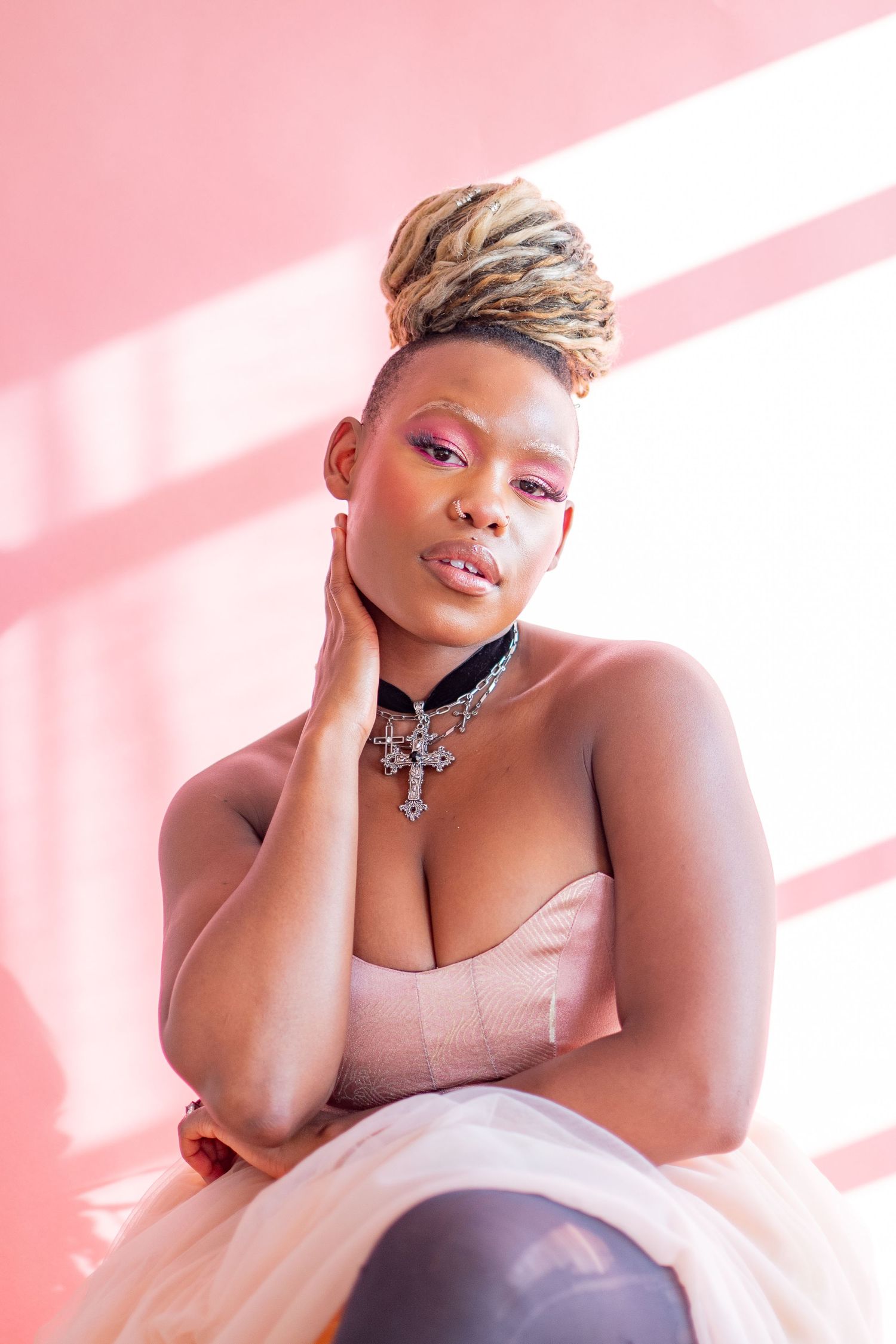 Portrait of a person with short blonde hair and pink makeup posing against a pink background with dramatic lighting.