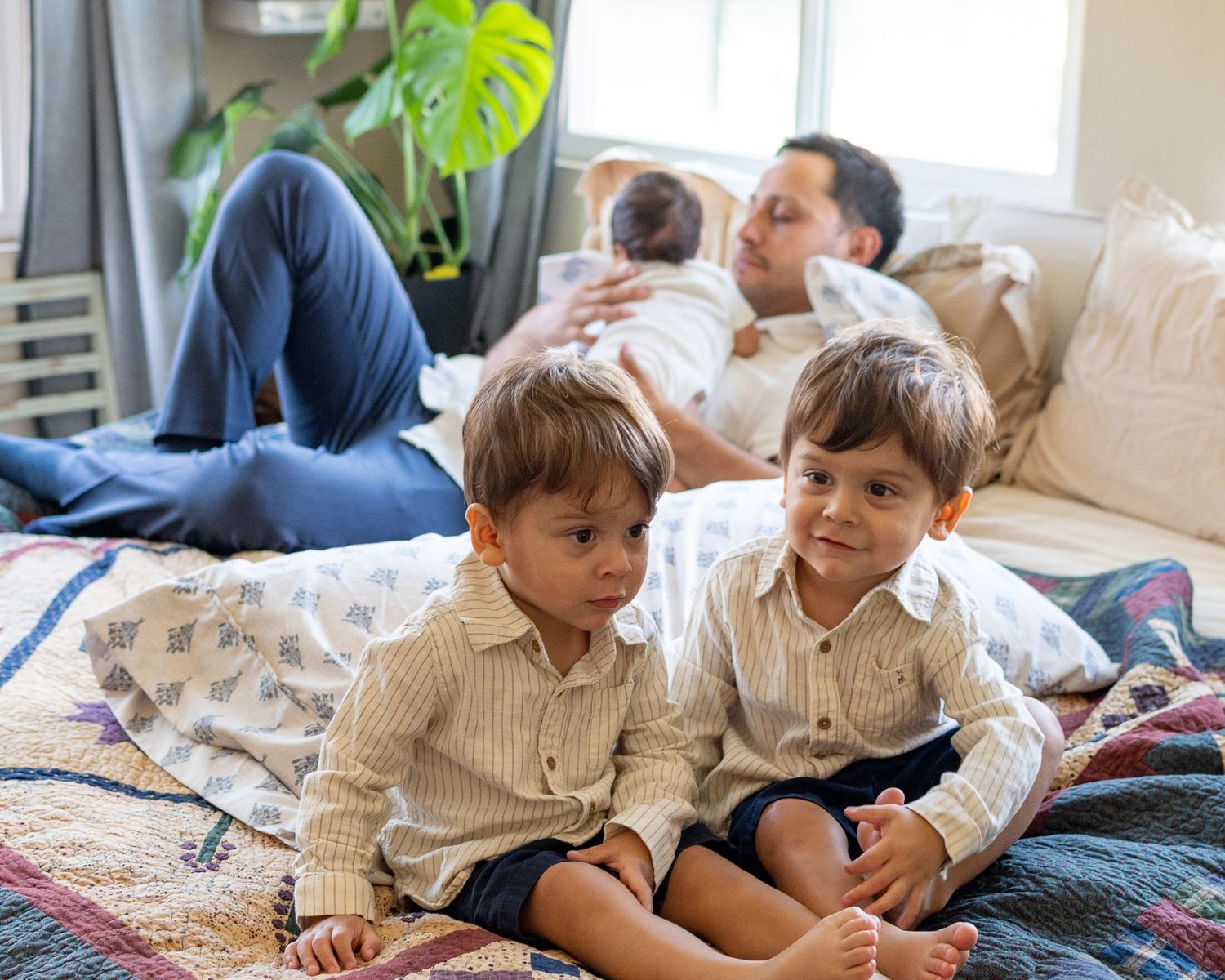 Two young boys sit on a bed while their father relaxes behind them holding a newborn baby.