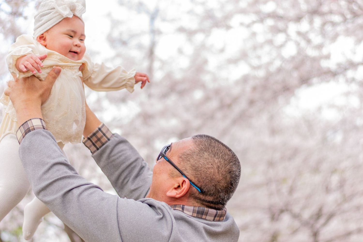 A person lifts a laughing baby wearing white clothing up in the air against a soft blurred background.