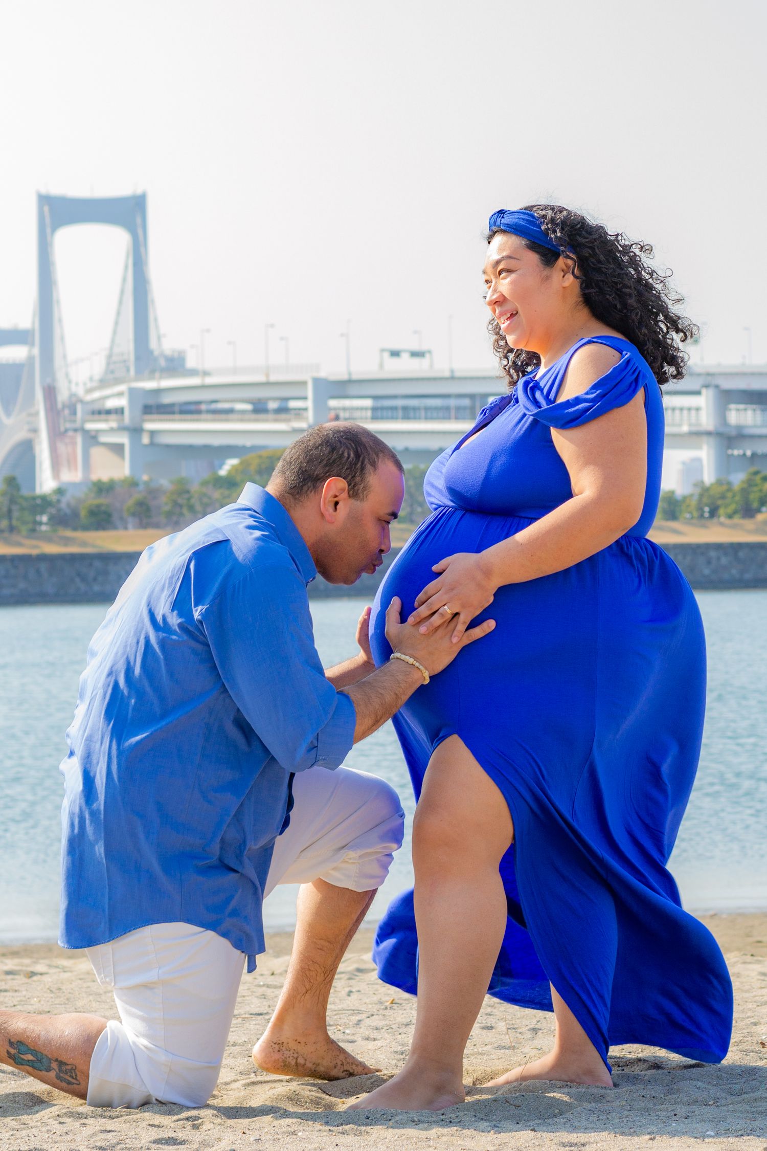 A couple in blue and white outfits share intimate moments on a sandy beach with a bridge in the background.
