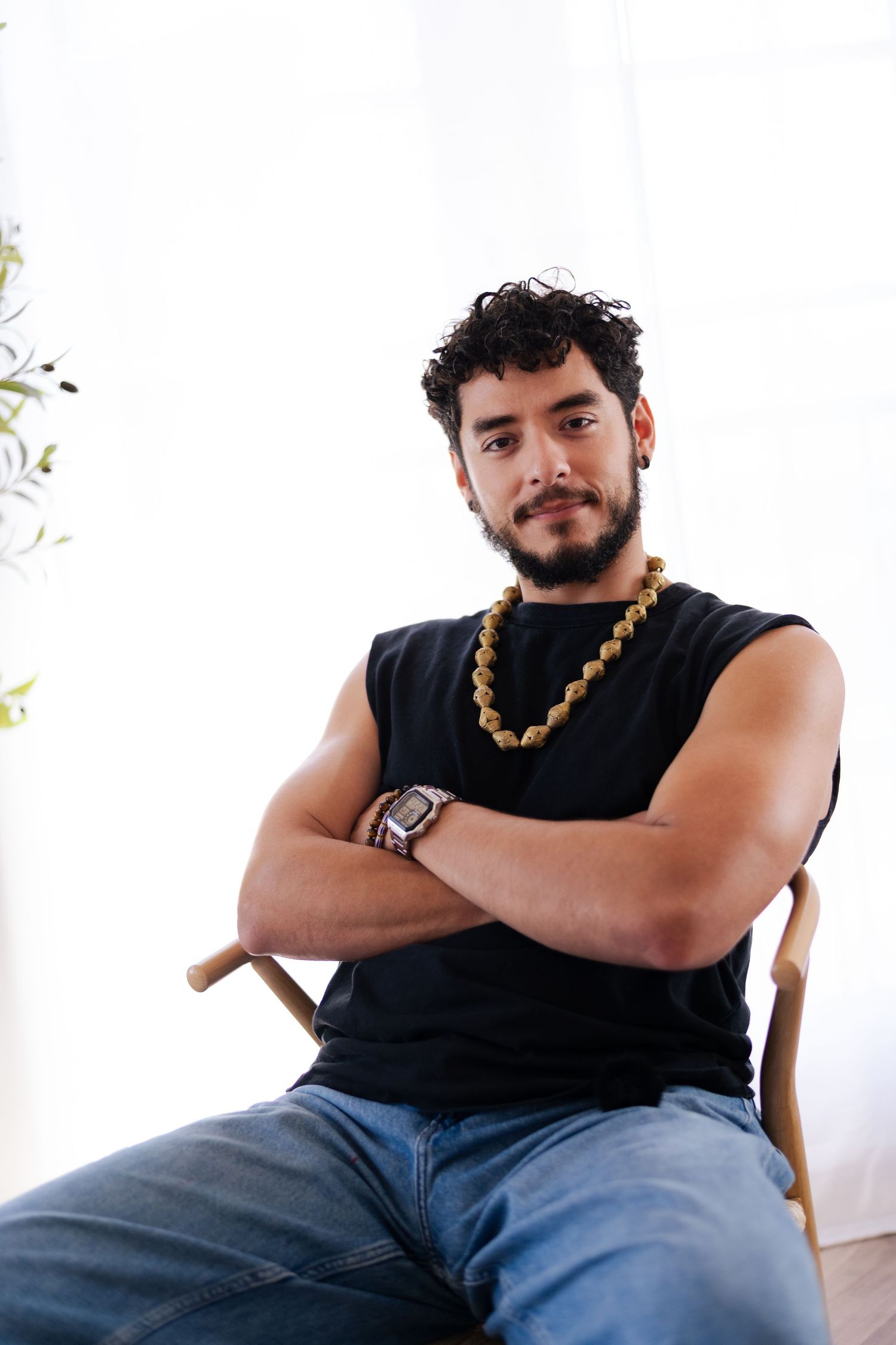 Person in black sleeveless shirt and jeans poses with crossed arms and beaded necklace against white background.
