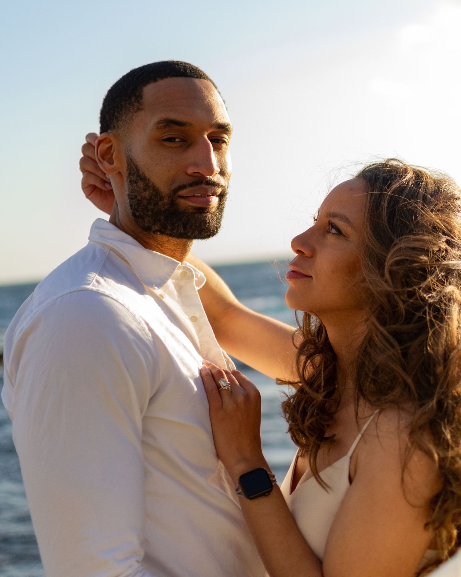A romantic couple embraces at sunset by the ocean, dressed in white and sharing an intimate moment.