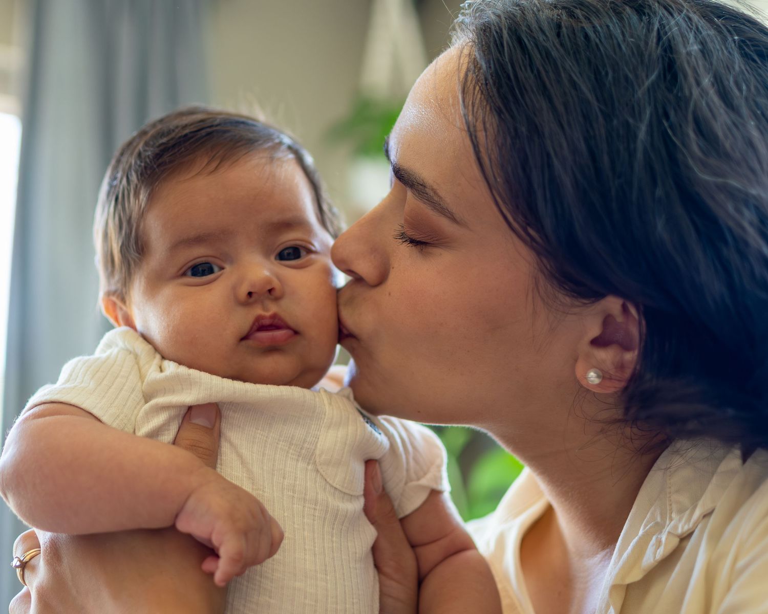 A mother tenderly kisses her adorable baby on the cheek while holding the infant in a bright, cozy room.